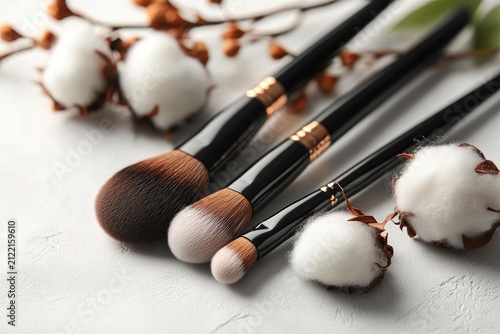 Cosmetic brushes with black handles lie near cotton flowers on a white surface. Perfect for beauty blogs, or skincare and make-up product promotions.
