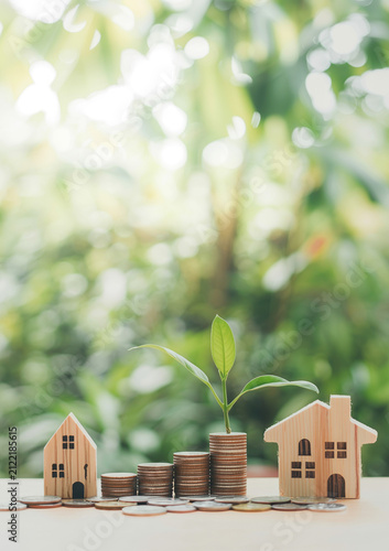 Small wooden houses and coin stacks with green sprout on wooden table.