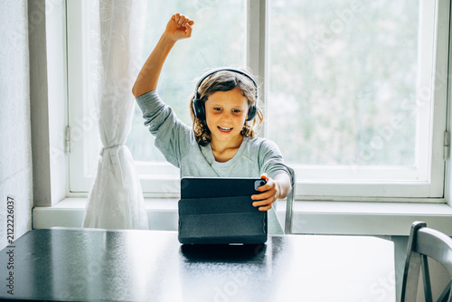 Happy long haired child boy  celebrating success while using tablet for online learning at home.