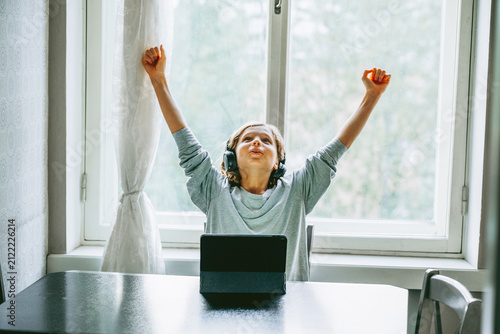 Young child wearing headphones raises both arms in celebration after achieving success on a digital tablet. Bright natural light and a comfortable home setting illustrate online learning.