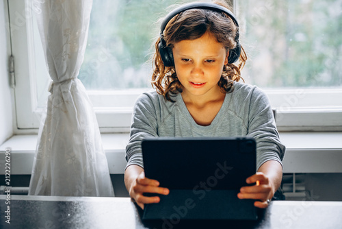 Focused long haired child boy using digital tablet with headphones during online learning at home.