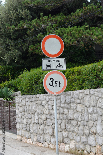 Картината върху платно A vertical stack of road signs showing a closed to all vehicles symbol with an exception panel in Croatian and a weight limit sign stands next to a stone wall