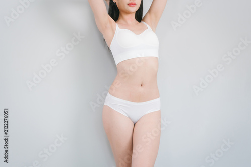 Young woman in white underwear standing against a plain gray wall with arms raised, showing a slim body and clean minimal background.