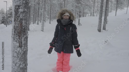 A happy child stands in a snow covered forest while heavy snow falls down upon her in winter. She wears a warm parka with a fur lined hood, colorful ski pants, and gloves outdoors