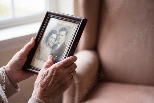 A close-up of an older person's wrinkled hands gently holding a framed black and white vintage photograph of a young couple by a window.