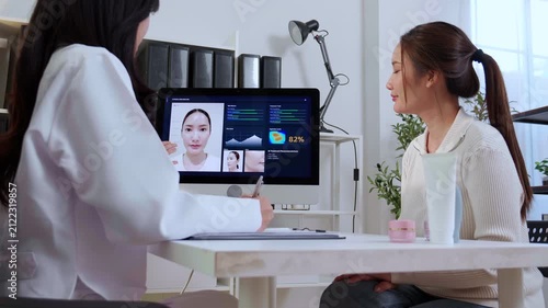 Asian woman doctor explaining ai face skin analysis on computer screen for female patient in clinic while dermatologist writing note on clipboard document, medical healthcare beauty consult.
