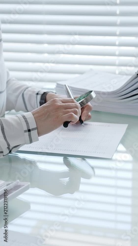 Financial professional performing calculations and writing data, preparing tax forms, managing budget, or completing administrative tasks on a bright office desk with stacks of documents