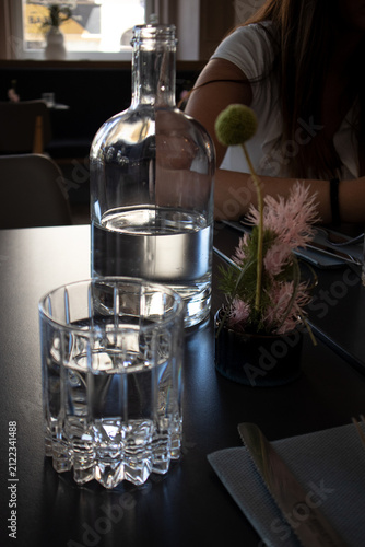 Bottiglia di vetro trasparente e bicchiere d'acqua riempiti sul tavolo scuro di un ristorante con piccoli fiori decorativi sullo sfondo