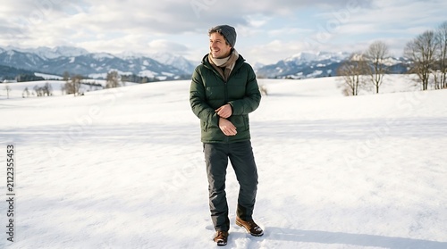 A young man wearing a warm winter jacket and beanie standing in a vast snowy field with mountains in the background during daytime