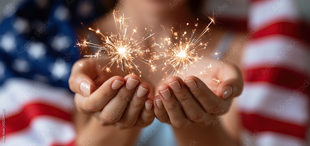 Obraz premium Festive summer scene with hands holding lit fireworks against a softly illuminated stars and stripes background.