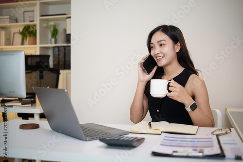 Smiling businesswoman talking on a smartphone while holding a coffee mug and working at her office desk.