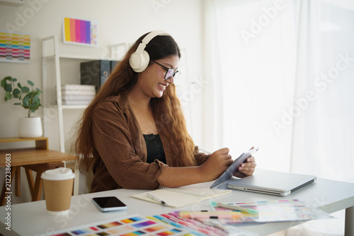 Smiling female designer wearing headphones writes while creating digital artwork while working remotely in a modern and organized workspace.