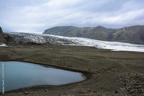 Vast Iceland landscape featuring a glacial tongue retreating from volcanic mountains, revealing a dark ground with a serene meltwater lake reflecting the cloudy sky