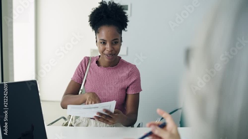 Young black woman checking in at a clinic, talking to the receptionist and signing a medical form before a doctor's appointment