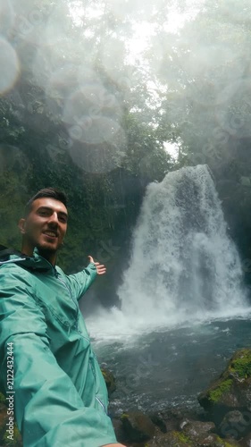 Young male content creator filming himself with a beautiful waterfall in the background at Arenal Volcano National Park, Costa Rica