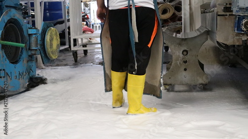 Lower body view of industrial worker standing in foamy wet floor conditions, wearing yellow rubber boots, black trousers with orange stripe, and protective apron inside busy manufacturing workshop