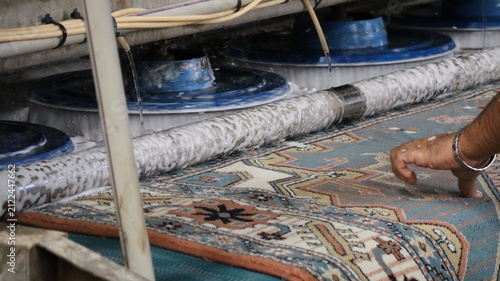 Close view of industrial carpet cleaning machine using rotating brushes and water jets to scrub patterned rug while technician hand guides fabric, with foam, hoses, metal pipe, and wet surface detail