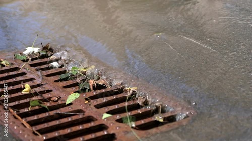 Detailed close up view of rainwater rushing through a rusty metal sewer drain grate covered in green leaves during a storm.