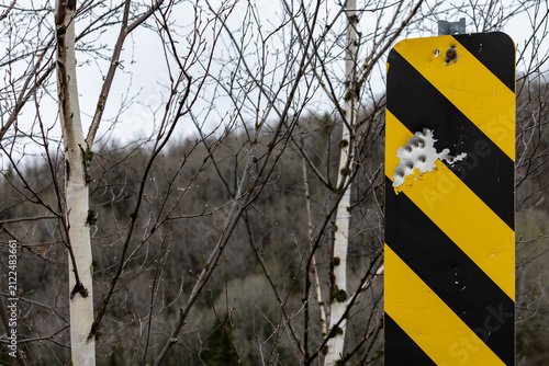 Obraz na płótnie Parents, Quebec, 19 May 2026 : A yellow and black striped warning sign shows multiple bullet holes in a natural, wooded environment