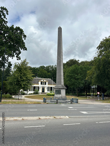 Bild auf Leinwand Stone obelisk monument De Naald in Apeldoorn next to a road, with a historic house and green trees in the background under a cloudy sky, Netherlands