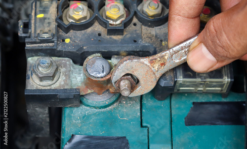 Close-up of an Asian man's hand tightening a car battery terminal bolt with an open-end wrench while servicing a vehicle battery to ensure a secure electrical connection.