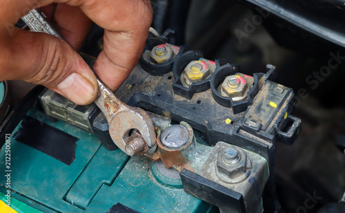 Close-up of an Asian man's hand tightening a car battery terminal bolt with an open-end wrench while servicing a vehicle battery to ensure a secure electrical connection.