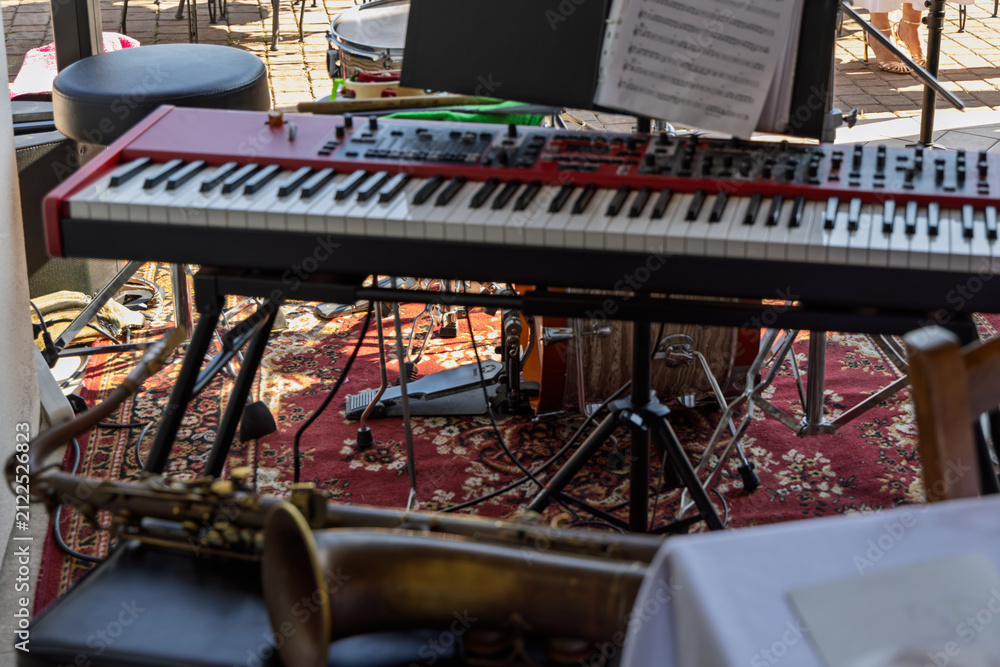 Fototapeta premium A red stage keyboard with sheet music leads a layered small-band setup, with a saxophone bell in front and an acoustic drum kit behind.