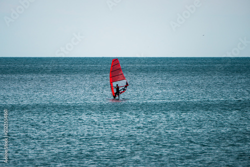 Obraz na płótnie A windsurfer glides across the calm sea with an orange sail under an overcast sky