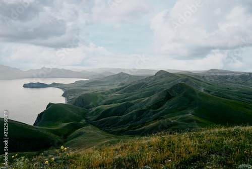 Bild auf Leinwand Expansive view of rolling green hills and rugged coastal cliffs overlooking a calm sea beneath a moody cloudy sky, evoking tranquility, remoteness and natural beauty in a serene summer landscape