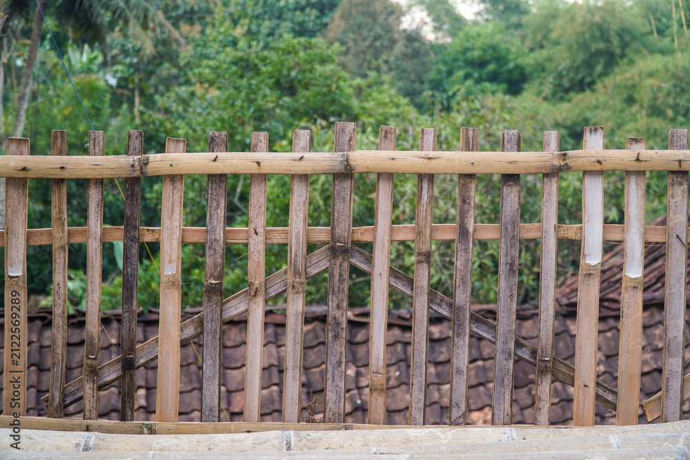 Fototapeta premium Rustic bamboo fence with weathered texture set against lush green forest backdrop and tiled roof. Natural, handmade construction ideal for rural or eco-friendly design projects.