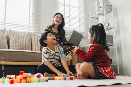 Asian mother working on laptop while her children play with building blocks at home
