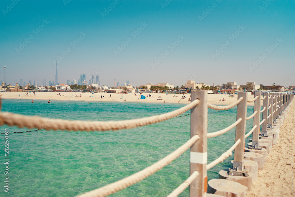 Fototapeta premium A scenic view of a beach with clear turquoise water, a wooden railing in the foreground, and a skyline featuring tall buildings in the background under a clear blue sky.