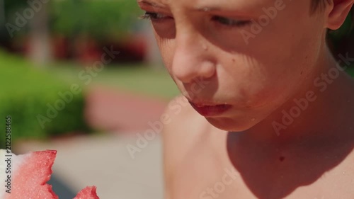 A close-up of a young boy taking a bite of a large, juicy slice of fresh watermelon on a sunny summer day. The vibrant red fruit, visible texture, and refreshing juice highlight healthy seasonal
