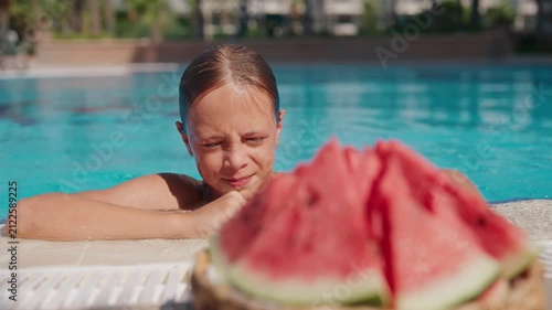 Young boy swimming toward a plate of fresh watermelon slices at an outdoor swimming pool on a sunny summer day. The juicy fruit is softly blurred in the foreground while the child is in sharp focus