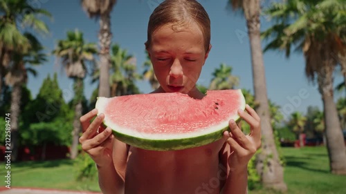 A happy young boy enjoys a large slice of fresh watermelon outdoors on a sunny summer day surrounded by palm trees and tropical greenery. Standing shirtless at a resort, he is about to take a bite of