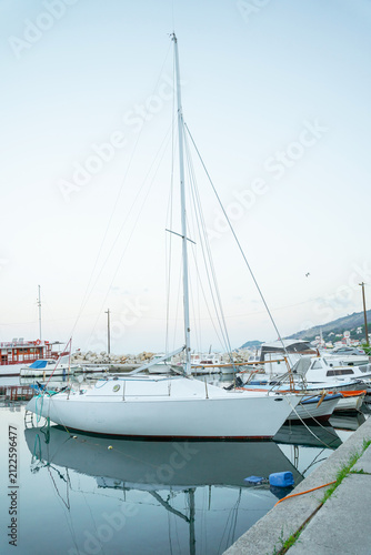 A white sailboat sits docked in a tranquil marina with its reflection visible on the still water surface against a clear sky.