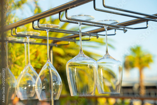 фотографія Clear wine glasses hanging from a metal rack at an outdoor bar with a sunny tropical background