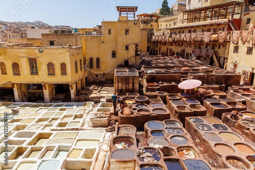 Fotografie An elevated view over the Chouara Tannery in the Fez medina, showing dozens of colorful stone dye pits arranged among traditional buildings, with workers moving between the vats