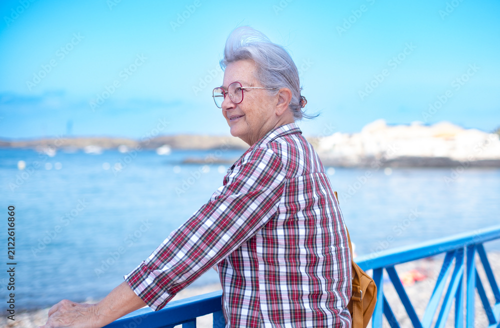 Fototapeta premium Senior adult woman enjoying a relaxing coastal vacation, smiling while looking at the calm blue sea, standing on a pier during retirement travel and leisure time