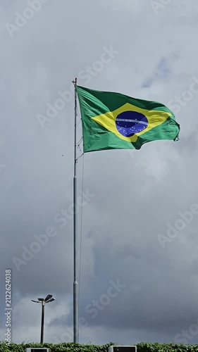 The Brazilian flag waves majestically on a flagpole against a dramatic, cloudy sky. The iconic green, yellow, and blue design is vibrant and striking