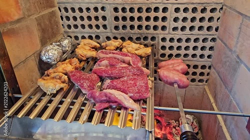 A delicious assortment of meats and chicken being grilled over an open flame. Raw steaks, sausages, and chicken pieces are arranged on a metal rack, with a baked potato and embers visible below