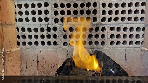 A fire burning in a brick fireplace with an artistic pattern of holes in the brickwork. The flames are bright orange and yellow, casting a warm glow on the dark logs and the surrounding bricks