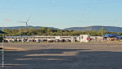 A wind farm with cars parked in the foreground and a wind turbine in the distance, under a clear sky. The scene depicts a roadside rural establishment with renewable energy in the background.