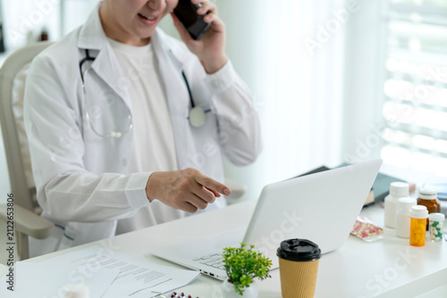 Male Doctor Using Laptop During Telemedicine Phone Consultation in Bright Modern Medical Office