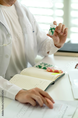 Male Doctor Holding Green Capsule Bottle Reviewing Medication Notes in Bright Medical Office