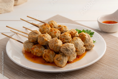 Charcoal grilled Thai Pork tendon meatballs on skewers on white plate with chilli sauce.
