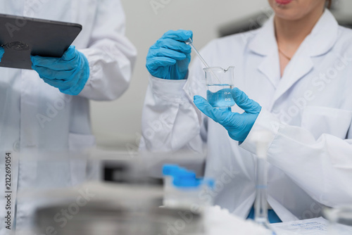 Professional female scientist using glass dropper with blue liquid in beaker for chemical analysis and medical research at modern laboratory, while colleague records data on digital tablet.