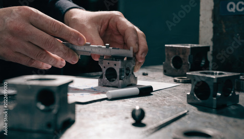 Hands using caliper to measure metal part, precision engineering, industrial workshop, mechanical component inspection, quality control, technical drawing visible, focused atmosphere