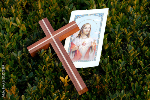 A wooden cross and an image of the Sacred Heart of Jesus rest side by side on a boxwood hedge with dense, green foliage.