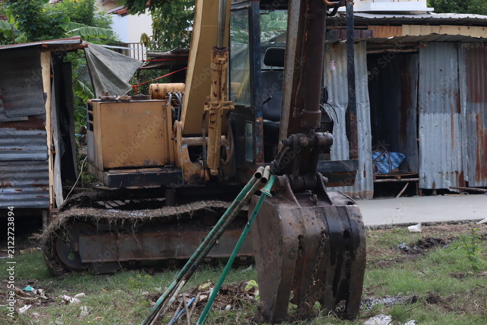 Fototapeta premium A heavy-duty yellow excavator parked on a grassy patch next to rustic corrugated metal shelters and debris.